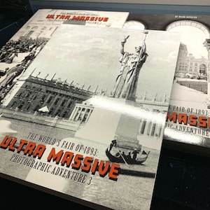 May include: A stack of books with the title "The World's Fair of 1893: Ultra Massive Photographic Adventure." The covers feature black and white historical images and orange text. The books are on a dark surface.