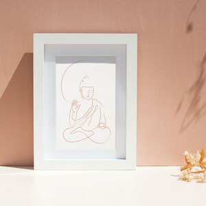 Puede incluir: Un marco blanco con un dibujo de l&iacute;nea de un Buda meditando en posici&oacute;n sentada. El dibujo es de color marr&oacute;n claro.