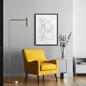 May include: A yellow armchair with black legs sits in a living room with a black and white line drawing of a person in a black frame on the wall, a gold floor lamp, a vase with pink flowers on a white side table, and a white entertainment center with a black TV.
