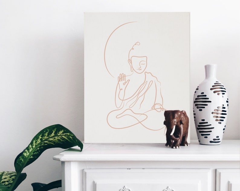 Puede incluir: Un dibujo de l&iacute;nea de un Buda meditando en un lienzo blanco con una figurita de elefante de madera marr&oacute;n y un jarr&oacute;n de cer&aacute;mica blanco con patrones geom&eacute;tricos negros en un estante blanco.