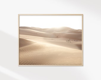 May include: A framed print featuring a desert landscape with rolling sand dunes. The image is bathed in warm sunlight, creating shadows and highlights on the textured sand. The artwork is in a light wooden frame.