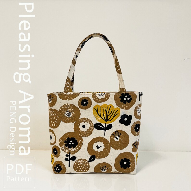 Op de afbeelding: Een witte tote bag met een bloemenpatroon in tinten bruin, geel en zwart. De tas heeft een bruin stoffen handvat en de tekst "Pleasing Aroma PDF Pattern" is afgedrukt aan de linkerkant van de afbeelding.