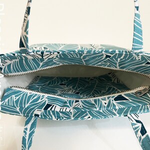 Op de afbeelding: Een teal tote bag met een wit bladmotief. De tas heeft een ritssluiting en twee bovenste handvatten. De tas is open en de binnenkant is gevoerd met witte stof. De tas heeft een klein ritsvak aan de binnenkant.