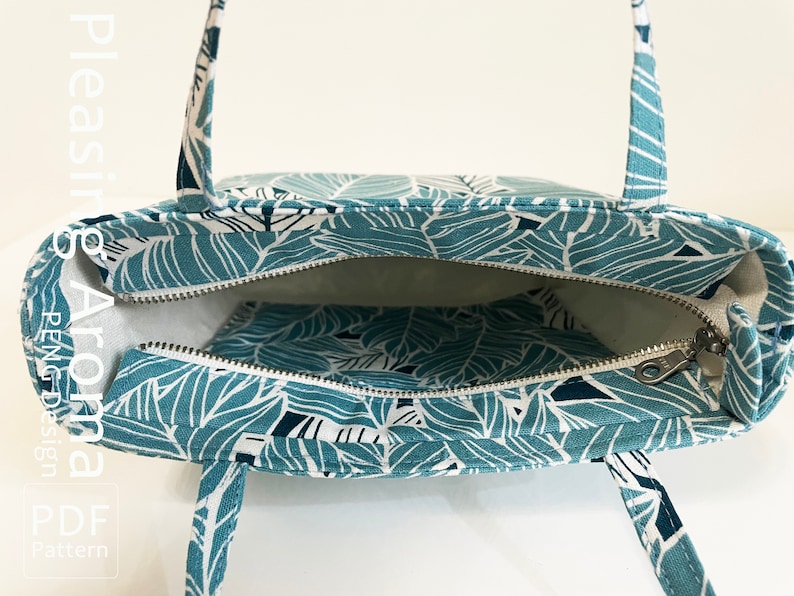 Op de afbeelding: Een teal tote bag met een wit bladmotief. De tas heeft een ritssluiting en twee bovenste handvatten. De tas is open en de binnenkant is gevoerd met witte stof. De tas heeft een klein ritsvak aan de binnenkant.