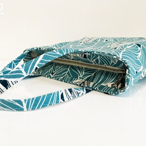 Op de afbeelding: Een tealblauwe tote bag met een wit en zwart bladmotief. De tas heeft een ritssluiting en een bovenste handvat.