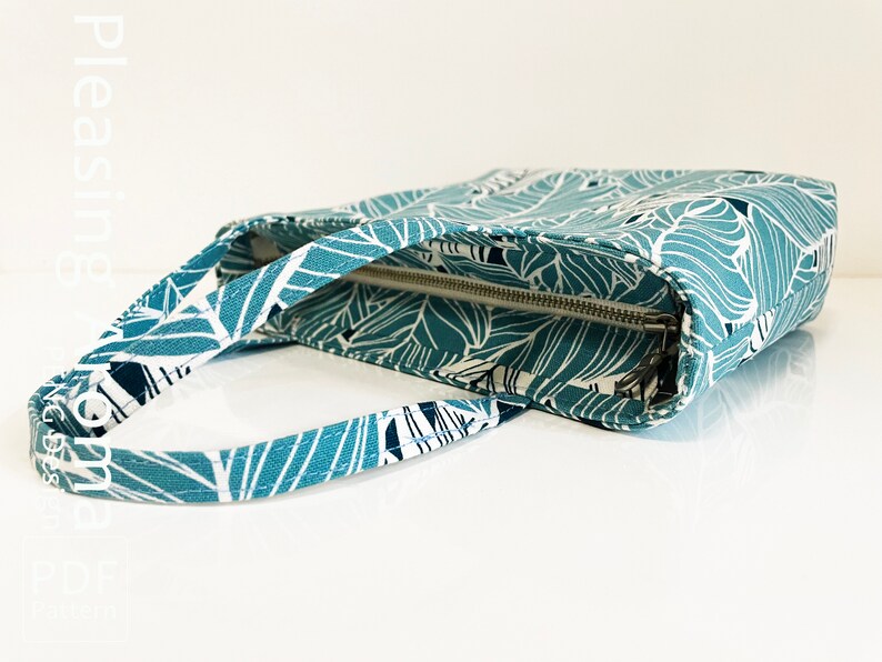 Op de afbeelding: Een tealblauwe tote bag met een wit en zwart bladmotief. De tas heeft een ritssluiting en een bovenste handvat.