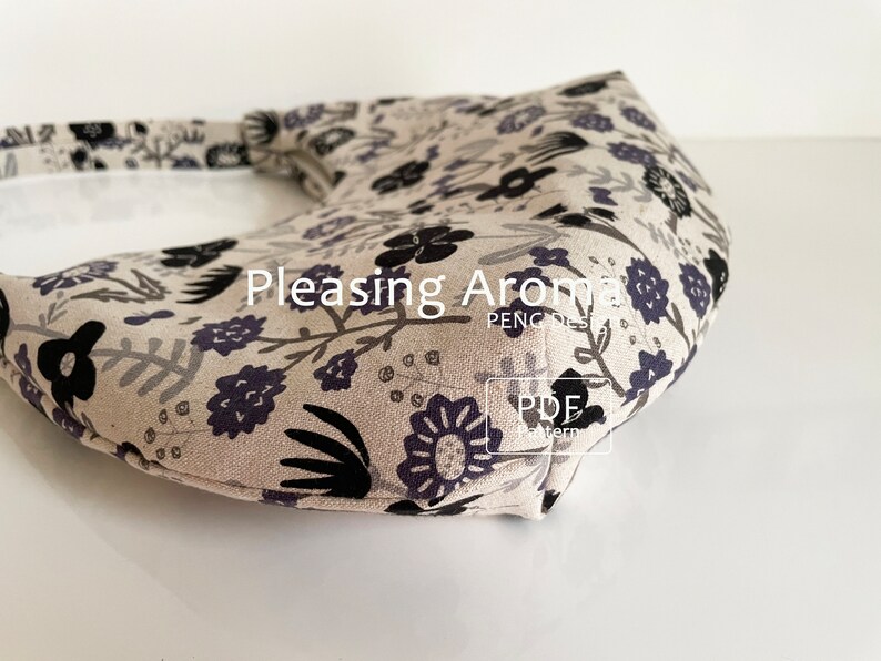 May include: A close-up of a beige fabric handbag with a black and purple floral print. The bag has a curved shape and a strap. The text "Pleasing Aroma" is visible on the bag.