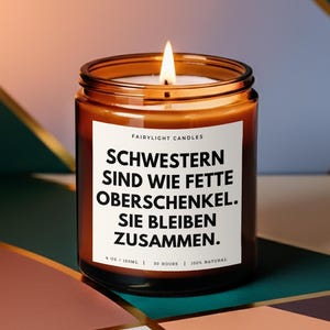 Könnte beinhalten: Eine brennende, bernsteinfarbene Glas-Kerze mit weißem Etikett. Auf dem Etikett steht "FAIRYLIGHT CANDLES" über dem Text "SCHWESTERN SIND WIE FETTE OBERSCHENKEL. SIE BLEIBEN ZUSAMMEN." Die Kerze ist 120ml und 100% natürlich.