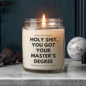 Könnte beinhalten: Eine brennende Sojawachskerze in einem Glas. Die Kerze hat ein weißes Etikett mit dem Text "HOLY SHIT, YOU GOT YOUR MASTER'S DEGREE" und "handmade with pure soywax". Der Hintergrund umfasst eine Marmorkugel und dunkle Dekoration.