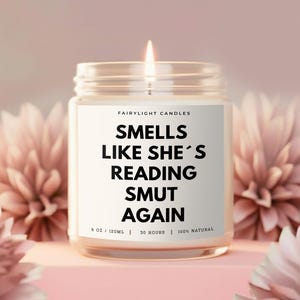 Könnte beinhalten: Eine brennende Kerze in einem Glas mit weißem Etikett. Auf dem Etikett steht "SMELLS LIKE SHE'S READING SMUT AGAIN". Die Kerze ist von rosa Blumen umgeben. Das Glas hat 120 ml und brennt 30 Stunden lang.