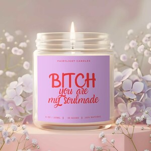 May include: A lit candle in a glass jar with a purple label. The label reads "BITCH you are my soulmade" in red text. The candle is surrounded by white and purple flowers, and sits on a pink pedestal.