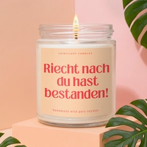 Könnte beinhalten: Eine Kerze im klaren Glas mit brennendem Docht und einem pfirsichfarbenen Etikett, das den Text "FAIRYLIGHT CANDLES" und "Riecht nach du hast bestanden!" in roter Schrift sowie "handmade with pure soywax" am unteren Rand zeigt.