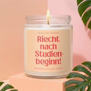 Könnte beinhalten: Eine Kerze im klaren Glas mit brennendem Docht, versehen mit einem beigen Etikett mit dem roten Text "FAIRYLIGHT CANDLES" und "Riecht nach Studien-beginn!", gefolgt von "handmade with pure soywax" in kleinerer Schriftgröße.