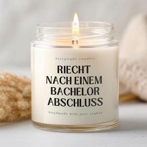 Könnte beinhalten: Eine brennende Kerze in einem transparenten Glas mit dem Text "RIECHT NACH EINEM BACHELOR ABSCHLUSS" in Schwarz. Die Kerze ist mit "fairylight candles" und "handmade with pure soywax" beschriftet. Der Hintergrund umfasst getrocknete Pflanzen und einen beigen Stoff.