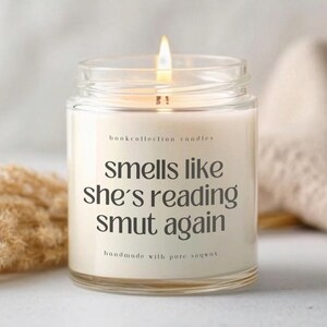 Puede incluir: Una vela encendida en un tarro de cristal transparente con el texto "smells like she's reading smut again." La vela est&aacute; etiquetada como "bookcollection candles" y "handmade with pure soywax."