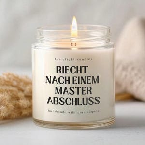 Könnte beinhalten: Eine brennende Kerze in einem transparenten Glasbehälter mit dem Text "RIECHT NACH EINEM MASTER ABSCHLUSS" in Schwarz. Die Kerze ist mit "fairylight candles" und "handmade with pure soywax" beschriftet. Der Hintergrund umfasst einen beigen Stoff und getrocknete Pflanzen.