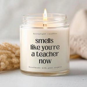 Könnte beinhalten: Eine brennende Kerze in einem transparenten Glas mit dem Text "smells like you're a teacher now". Die Kerze ist mit "fairylight candles" und "handmade with pure soywax" beschriftet. Der Hintergrund umfasst getrocknete Pflanzen und einen beigen Stoff.