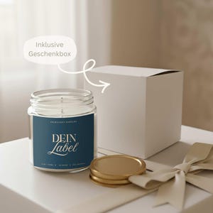Puede incluir: Una vela en tarro de cristal transparente con una etiqueta azul que dice "DEIN Label" en letras doradas. La vela, con tapa dorada, est&aacute; junto a una caja de regalo blanca y una cinta color crema. La etiqueta dice "Fairylight Candles".