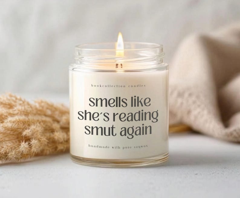 Puede incluir: Una vela encendida en un tarro de cristal transparente con el texto "smells like she's reading smut again." La vela est&aacute; etiquetada como "bookcollection candles" y "handmade with pure soywax."