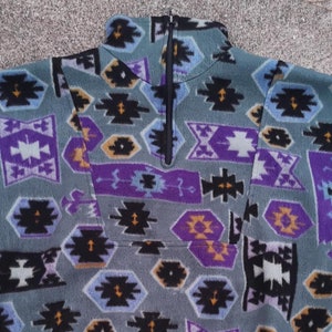 Puede incluir: Un jersey de polar verde, morado, negro y amarillo con un patrón geométrico. El jersey tiene una cremallera en la parte delantera y un cuello alto.