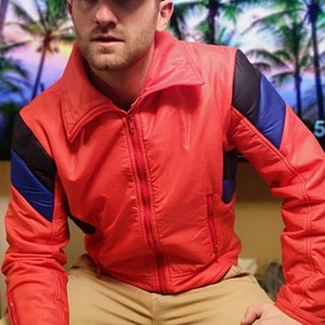 Könnte beinhalten: Ein Mann trägt eine rot-blaue Skijacke mit einer schwarzen Baseballkappe. Die Jacke hat einen Reißverschluss und einen hohen Kragen. Der Mann sitzt mit gekreuzten Beinen. Der Hintergrund ist ein verschwommenes Bild eines tropischen Strandes.