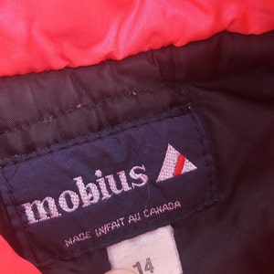 Pode incluir: Um r&oacute;tulo azul com a palavra "mobius" em letras brancas e um logotipo triangular vermelho e branco. O r&oacute;tulo tamb&eacute;m diz "Made in/fait au Canada".