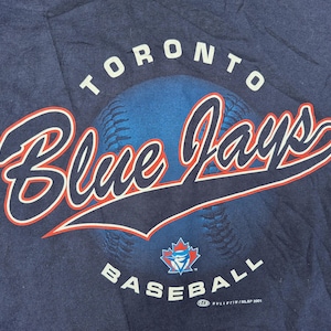 May include: Navy blue t-shirt with the Toronto Blue Jays logo. The design includes the words "TORONTO" and "BASEBALL" in white, with "Blue Jays" in a stylized script with red and white accents. A baseball graphic is in the background.