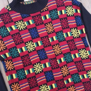 Può includere: Un maglione colorato con un motivo geometrico di quadrati rossi, neri, gialli, verdi e blu con un disegno a fiocco di neve.