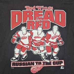 May include: Black t-shirt with a red and white graphic of three hockey players. The text "Dread the Red" and "Russian to the Cup" are in red. A logo of a winged wheel is at the bottom of the shirt.