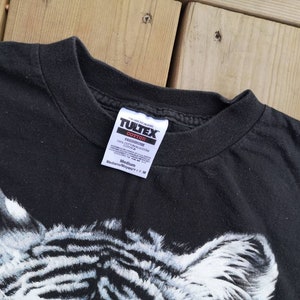 Może przedstawiać: Czarny T-shirt z białym nadrukiem zebry. Koszulka ma metkę Tultex z napisem "Preshrunk 100% Cotton" i "Medium/Moyen/M".