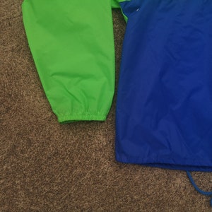 K&ouml;nnte beinhalten: Nahaufnahme einer leuchtend gr&uuml;nen und blauen Jacke. Die Jacke hat einen Kordelzug am unteren Saum und elastische B&uuml;ndchen. Die Jacke besteht aus einem leichten, m&ouml;glicherweise Nylon-Material. Die Farben sind lebendig und die Jacke scheint neu zu sein.