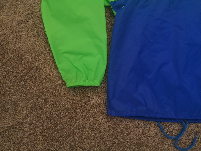 K&ouml;nnte beinhalten: Nahaufnahme einer leuchtend gr&uuml;nen und blauen Jacke. Die Jacke hat einen Kordelzug am unteren Saum und elastische B&uuml;ndchen. Die Jacke besteht aus einem leichten, m&ouml;glicherweise Nylon-Material. Die Farben sind lebendig und die Jacke scheint neu zu sein.