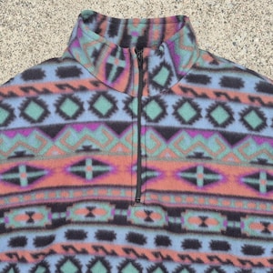 May include: A patterned fleece pullover in blue, orange, teal, purple, and black. The geometric design features diamond and cross shapes. It has a high collar and a black zip.