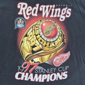 May include: Black t-shirt with a gold championship ring design, the text reads "1997 Stanley Cup Champions." The ring has a red centre with a logo and is surrounded by diamonds. The words "Red Wings" are at the top.