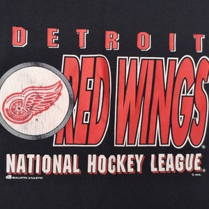 May include: Black t-shirt featuring a red and white Detroit Red Wings logo and the text "DETROIT RED WINGS NATIONAL HOCKEY LEAGUE".