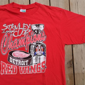 May include: Red t-shirt featuring a graphic of the 1997 Stanley Cup Champions Detroit Red Wings. The graphic includes the Stanley Cup trophy, a hockey puck with the Red Wings logo, and the text "Stanley Cup Champions 1997 Detroit Red Wings".