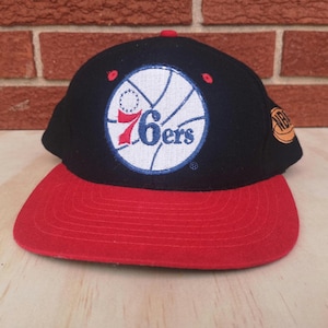 Op de afbeelding: Zwarte en rode baseballpet met een wit en blauw Philadelphia 76ers logo. Het logo toont een basketbal met de teamnaam en een ster.
