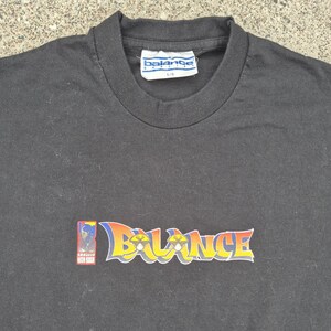 Könnte beinhalten: Schwarzes T-Shirt mit Rundhalsausschnitt und dem Wort "Balance" in einer farbenfrohen Schriftart im Graffiti-Stil auf der Vorderseite. Das Shirt hat auch eine kleine Grafik auf der linken Seite des Wortes.