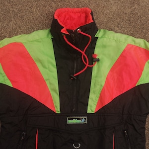 Pode incluir: Uma jaqueta de esqui preta, verde e vermelha com fecho de correr e capuz com cord&atilde;o. A jaqueta tem um logotipo que diz "Mountain" na frente.