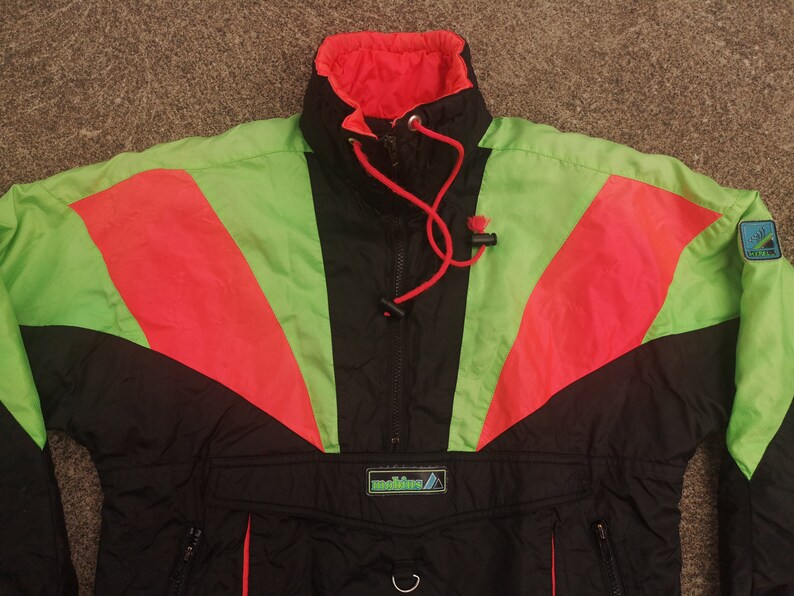 Pode incluir: Uma jaqueta de esqui preta, verde e vermelha com fecho de correr e capuz com cord&atilde;o. A jaqueta tem um logotipo que diz "Mountain" na frente.