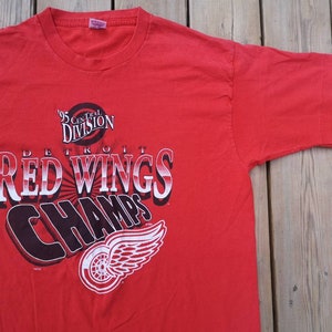 May include: Red t-shirt with a white and black graphic of the Detroit Red Wings logo and the text "'95 Central Division Detroit Red Wings Champs".