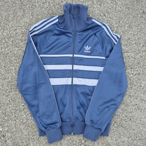 Könnte beinhalten: Blaue Trainingsjacke mit weißen Streifen und dem Adidas-Logo. Die Jacke hat einen durchgehenden Reißverschluss, einen hohen Kragen und gerippte Bündchen. Die Jacke ist aus glänzendem, sportlichem Material.