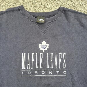 May include: A navy blue sweatshirt with the words "MAPLE LEAFS" in white, and a white maple leaf logo. Below the text is the word "TORONTO". The sweatshirt has a crew neck and a small tag at the back of the neck.