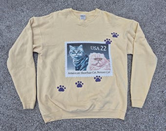 May include: A light yellow sweatshirt featuring a postage stamp design with two cat portraits and the text "USA 22". The sweatshirt also has purple paw prints scattered across the front.