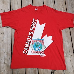 May include: Red t-shirt featuring a white Canadian flag design. The flag has the text "Canada's First" and "Toronto Blue Jays World Series 1992 Champions" printed on it.