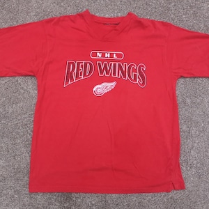May include: Red t-shirt featuring the NHL Red Wings logo and text. The logo is a red wing with a hockey puck in the centre. The text is in white with a red outline.
