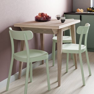 May include: A light green wooden dining table with four light green chairs. The table is set with a bowl of fruit and two mugs. The table is in a kitchen with light green cabinets and a pink wall.