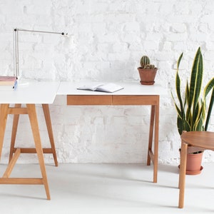 May include: A white desk with a wooden frame and two drawers. The desk has a corner extension and is set against a white brick wall. A wooden chair with a blue seat cushion sits next to the desk. A potted plant and a lamp are also visible.