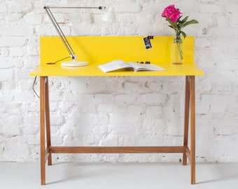May include: A yellow desk with a wooden frame and a lamp on the top. The desk is against a white brick wall and has a flower in a glass vase on the right side.