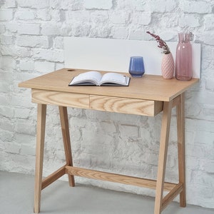 Peut inclure: Un bureau en bois clair avec deux tiroirs et un dosseret blanc. Le bureau a un design simple avec des pieds inclinés et une petite étagère en dessous. Il y a un livre, un verre et deux vases avec des fleurs sur le bureau.